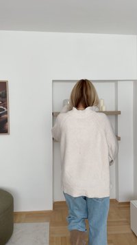 Pulover tricotat de culoare bej deschis, cu guler ribat, purtat împreună cu blugi relaxați, de culoare albastră deschisă. Pe rafturile din fundal se află lumânări și obiecte din sticlă.