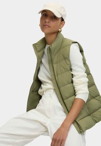 Kaki puffer vest met hoge collar en ritssluiting, gelaagd over een wit long-sleeve shirt. Witte canvas broek. Lichtgekleurde pet.