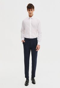 Biała koszula z długim rękawem z zapięciem na guziki, zestawiona z granatowymi spodniami o teksturowanym wzorze. Model stoi w czarnych butach eleganckich.