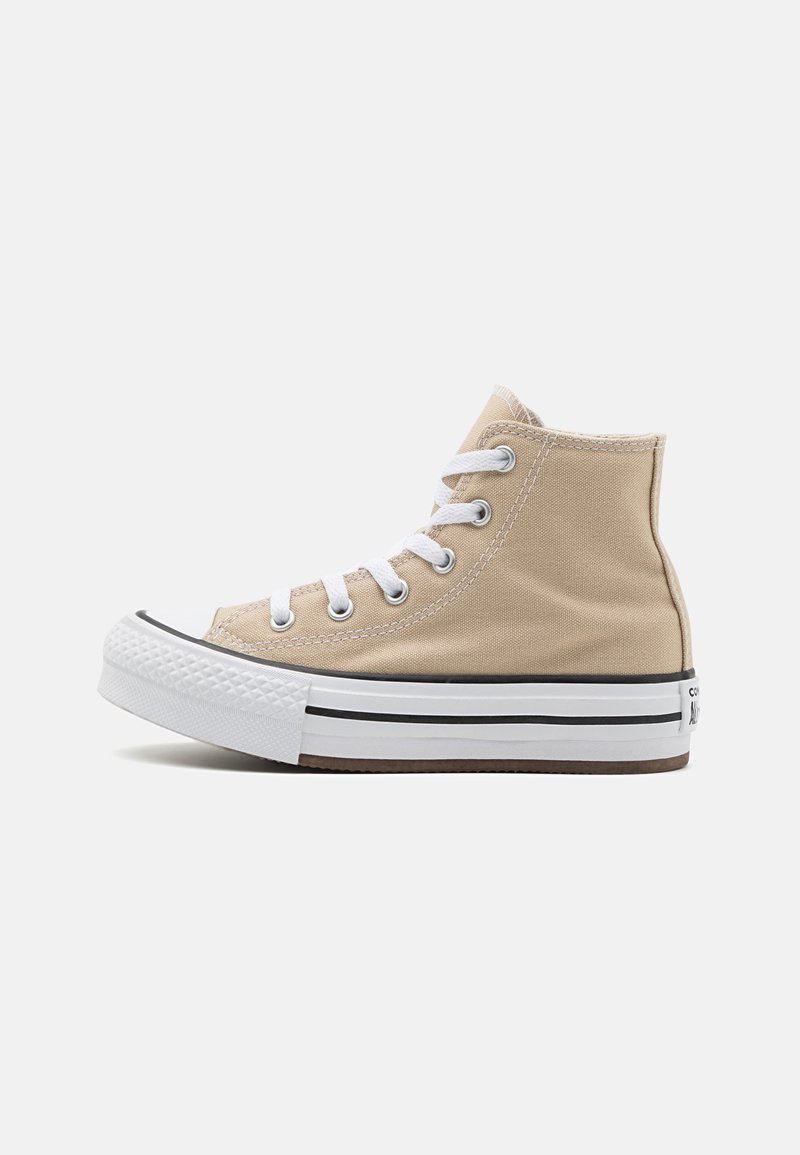 Zapatilla de lona de caña alta beige con cordones blancos, puntera de goma y rayas negras en la suela media. Presenta una superficie texturizada y un branding mínimo.