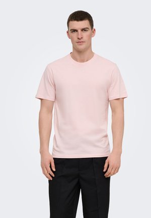 Young man wearing a light pink short-sleeve T-shirt and black pants, standing against a plain light background.