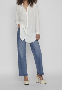 Chemisier blanc boutonné à manches longues, associé à un jean en denim bleu à taille haute et jambes larges, accompagné de chaussures crème à talons carrés.