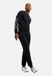 Sort sweatshirt og joggers i tykt stof, med et logo på brystet og tekst på ærmet, parret med hvide sneakers.