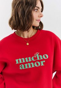 Czerwona bluza z napisem "mucho amor" w zielonym kolorze. Materiał wydaje się miękki, a fason ma luźny okrągły dekolt.