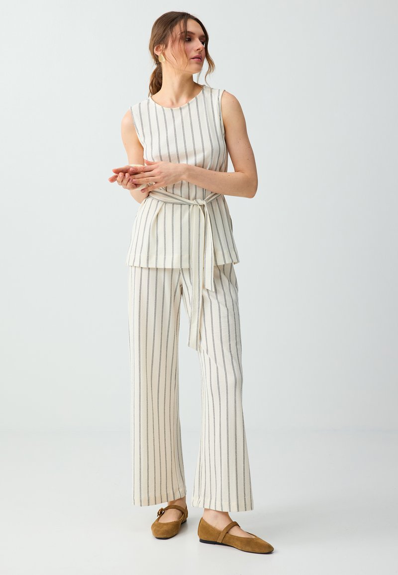 Femme portant un haut blanc sans manches et un pantalon large à fines rayures noires verticales, ceinture nouée et chaussures plates marron, debout sur fond blanc.
