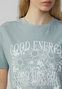 Lichtblauwe katoen t-shirt met een bloemenontwerp en witte tekst die luidt: "GOEDE ENERGIE" en "GOEDE GEDACHTEN WORDEN GOEDE DINGEN."