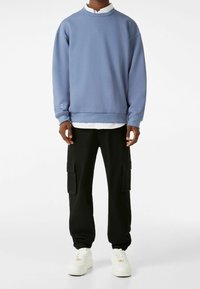 Felpa blu con polsini e orlo a costine, abbinata a pantaloni cargo neri con tasche laterali e polsini elastici. Sneakers bianche completano il look.