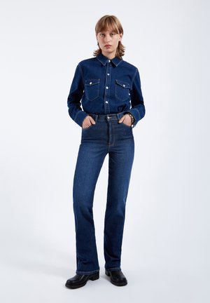 Person, der står iført mørkeblå denimskjorte og højtsiddende jeans med sorte ankelstøvler på en ensfarvet baggrund.