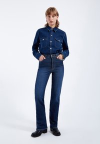 Camicia in denim scuro con bottoni e due tasche sul petto, abbinata a jeans a vita alta a campana, entrambi di un blu intenso. Indossata con zoccoli neri.
