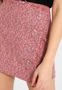 Mini jupe en tissu texturé rouge et blanc, avec des détails en franges et quatre boutons ronds argentés sur le devant.