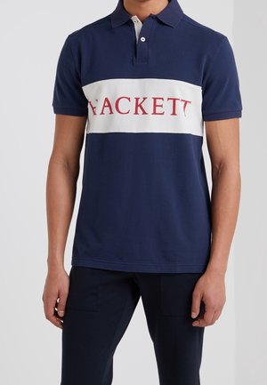 Homme portant un polo bleu marine avec une bande blanche horizontale et le texte rouge "HACKETT" sur la poitrine, associé à un pantalon foncé.