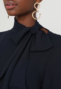 Nainen, joka pukeutuu suurilla kultaisilla geometrisilla korviksilla ja laivastonsiniseen paitapuseroon, jossa on solmittu rusetti kaulassa.