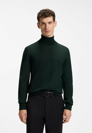 Uomo con capelli corti e ricci che indossa un maglione a collo alto verde scuro e pantaloni neri, in piedi davanti a uno sfondo bianco uniforme.