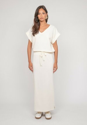 Femme aux longs cheveux bruns portant une robe blanche à col en V avec taille à cordon de serrage et des baskets blanches, debout devant un fond clair uni.