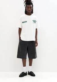 White t-shirt with green graphics, paired with dark gray knee-length shorts. Black shoes and white crew socks complete the outfit.
