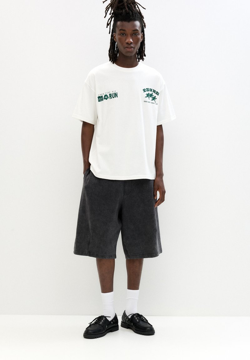 Hvid t-shirt med grøn grafik, parret med mørkegrå knælange shorts. Sorte sko og hvide ankelstrømper fuldender outfittet.