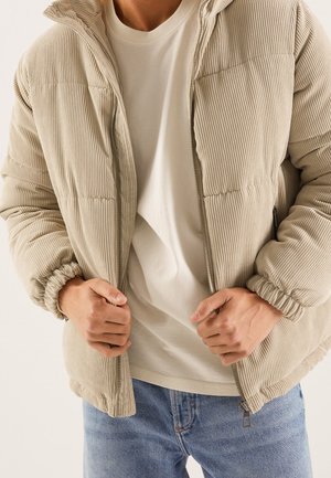 Personne portant une doudoune en velours côtelé beige par-dessus une chemise blanche et un jean bleu, les mains serrant la fermeture éclair de la veste près de la taille.
