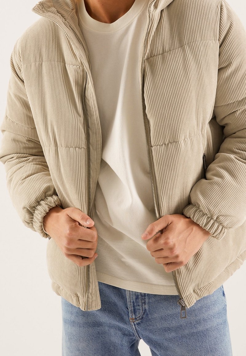 Personne portant une doudoune en velours côtelé beige par-dessus une chemise blanche et un jean bleu, les mains serrant la fermeture éclair de la veste près de la taille.