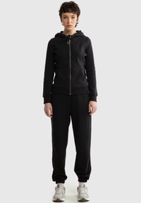 Jonge vrouw staand, gekleed in een zwarte hoodie met rits, zwarte joggingbroek en witte sneakers, op een effen lichtgrijze achtergrond.