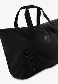 Bolsa de deporte negra hecha de tela texturizada, con dos asas, una correa ajustable y un parche de logo prominente en la parte frontal. Cuenta con un clip.
