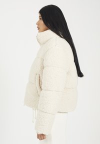 Veste rembourrée de couleur crème en fausse fourrure moelleuse. Dotée d'un col montant, d'épaules tombantes et d'un ourlet ajustable avec cordon.