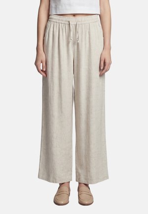 Persoon draagt losse, lichtbeige linnen broek met elastische tailleband en trekkoord, gecombineerd met beige instapschoenen en een wit cropped top.