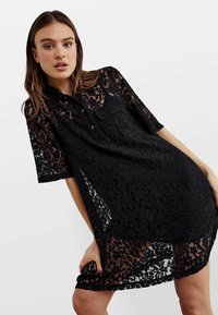 Rochie neagră din dantelă, cu mâneci scurte și guler cu nasturi. Prezintă un model floral și o textură ușor transparentă, tăietură până la genunchi.