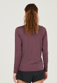 Langärmliges Hemd aus dunkellila Stoff, mit Rundhalsausschnitt und glatter Textur. Sichtbare Nähte am Saum und an den Bündchen.
