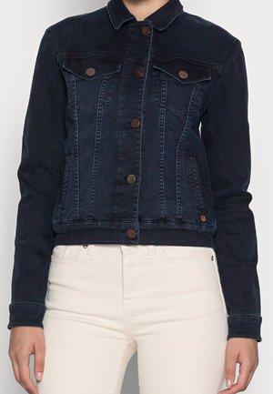 Osoba nosící tmavě modrou džínovou bundu s měděnými knoflíky a bílé kalhoty, zobrazená od ramen po boky.