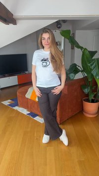 Γυναίκα με μακριά μαλλιά στέκεται μέσα στο σπίτι, φοράει λευκό μπλουζάκι με γραφικό γάτας, μαύρο παντελόνι, λευκά αθλητικά παπούτσια, κοντά σε πορτοκαλί καναπέ και μεγάλο γλαστράκι.