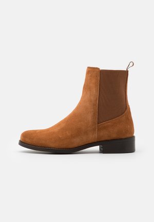 Brązowy zamszowy but Chelsea z elastycznym panelem bocznym i pętelką do zakładania na czarnej podeszwie i niskim obcasie na białym tle.