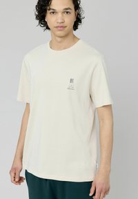 T-shirt crème à manches courtes avec un col rond, présentant un petit motif graphique noir sur la poitrine. Texture en coton douce.