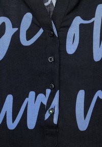 Ciemnoniebieska koszula z dużym wzorem tekstowym w jasnoniebieskim kolorze. Posiada kołnierzyk, zapięcie na guziki z przodu oraz zielony akcent guzikowy. Tekst ma nieregularny kształt.