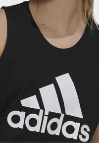 Svart bomullstopp utan ärmar med en stor vit Adidas-logotyp, rund halsringning och en stilren design som framhäver rena linjer och en slät textur.