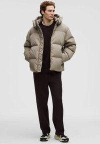 Mann trägt eine beige gesteppte Jacke mit Kapuze, schwarzen Pullover, schwarze Hose und schwarz-weiße Turnschuhe, steht mit den Händen in den Taschen.