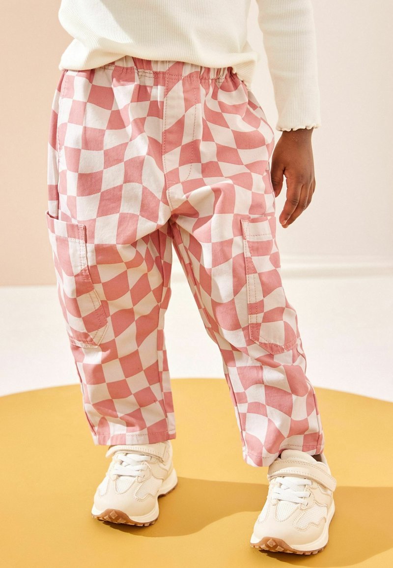 Enfant portant un pantalon à carreaux roses et blancs avec des poches cargo, des baskets blanches et un haut blanc à manches longues, debout sur un sol jaune.