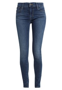 Jeans ajustados de color azul oscuro con botón frontal, cremallera, pasa cinturón y bolsillos delanteros y traseros, exhibidos sobre un fondo blanco liso.