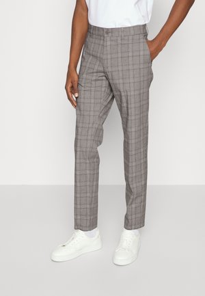 Mand iført grå ternede bukser, hvide sokker, hvide sneakers og en hvid skjorte med den ene hånd i lommen mod en ensfarvet baggrund.
