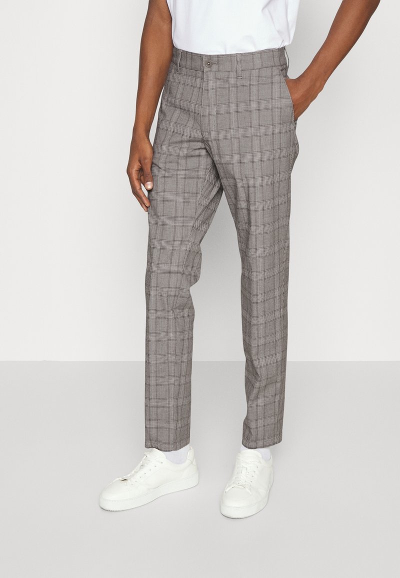 Homme portant un pantalon à carreaux gris, des chaussettes blanches, des baskets blanches et une chemise blanche, une main dans la poche, sur un fond uni.
