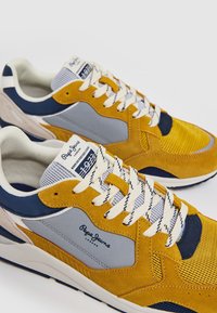 Sneakers gialli e blu navy realizzati in materiali scamosciati e mesh, con un pannello grigio, accenti beige, superfici testurizzate e lacci con motivo.