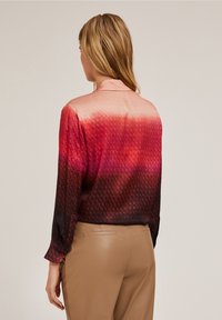 Blus i röd och rosa gradient med en strukturerad mönster, långa ärmar och krage bak, i kombination med åtsittande beige byxor.