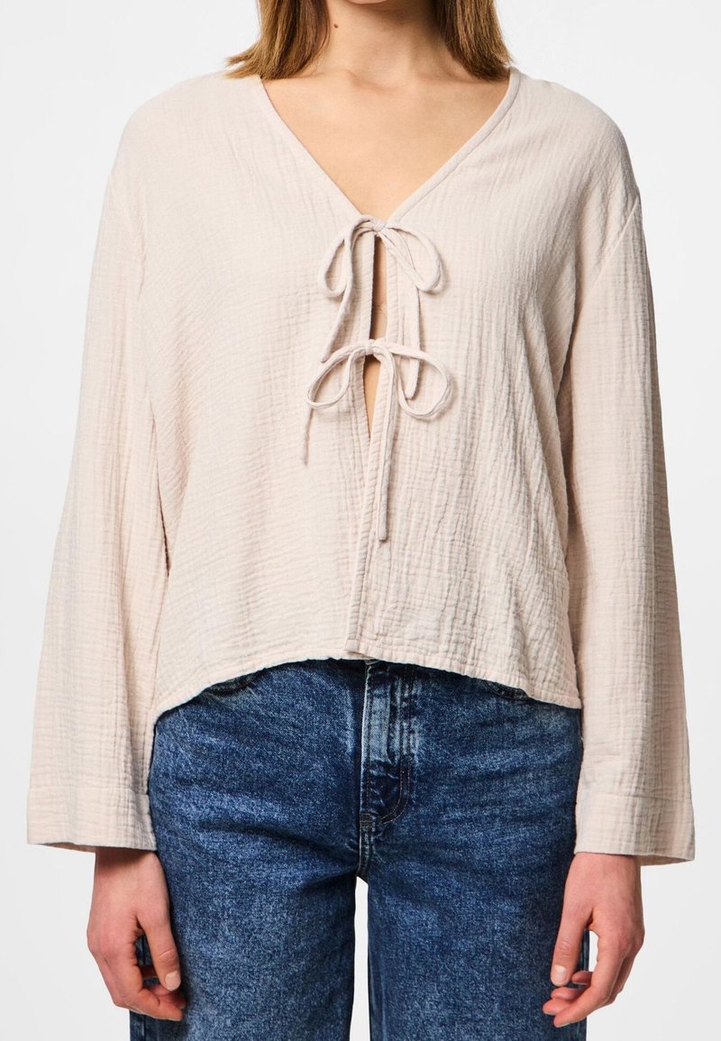 Blusa beige chiaro, strutturata, con scollatura a V e dettaglio di fiocco frontale. Presenta maniche lunghe e ampie e un orlo corto, abbinata a jeans blu scuro.