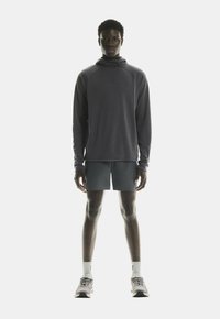Sivý dlhý rukáv športový vrh s kapucňou, skombinovaný s tmavými šortkami. Model má na sebe svetlé tenisky a pruhované ponožky. Minimalistický dizajn.
