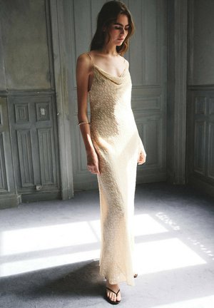 Femme en robe maxi sans manches texturée crème et sandales noires, debout dans une pièce ensoleillée avec des murs lambrissés gris.