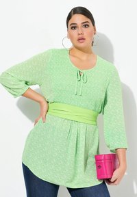 Blouse verte à motifs avec des manches trois-quarts, détail à nouer au cou et taille froncée. Assortie à un jean foncé et à une pochette rose.