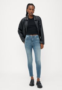 Velika jakna iz umetnega usnja v črni barvi, črn ribast crop top, svetlo modri skinny jeans, črne natikače, ravni lasje z rahlo teksturo.