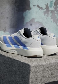 Scarpe da corsa Adidas bianche e blu con suole Lightstrike Pro posizionate su una superficie di pietra contro uno sfondo di muro strutturato.