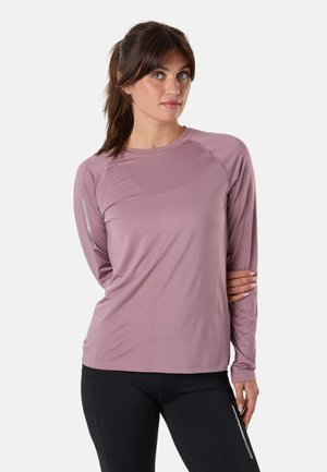Femme aux cheveux bruns attachés en queue de cheval, portant un haut de sport mauve à manches longues et des leggings noirs avec des détails réfléchissants, debout sur fond blanc.