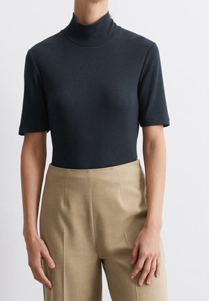 Person mit enganliegendem schwarzen Kurzarm-Rollkragenpullover und hoch taillierter beigefarbener Hose, stehend vor hellem, schlichten Hintergrund.