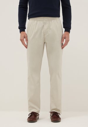 Man die beige elastische broek met tailleband, marineblauw shirt met lange mouwen en bruine leren veterschoenen draagt, staand tegen een effen achtergrond.